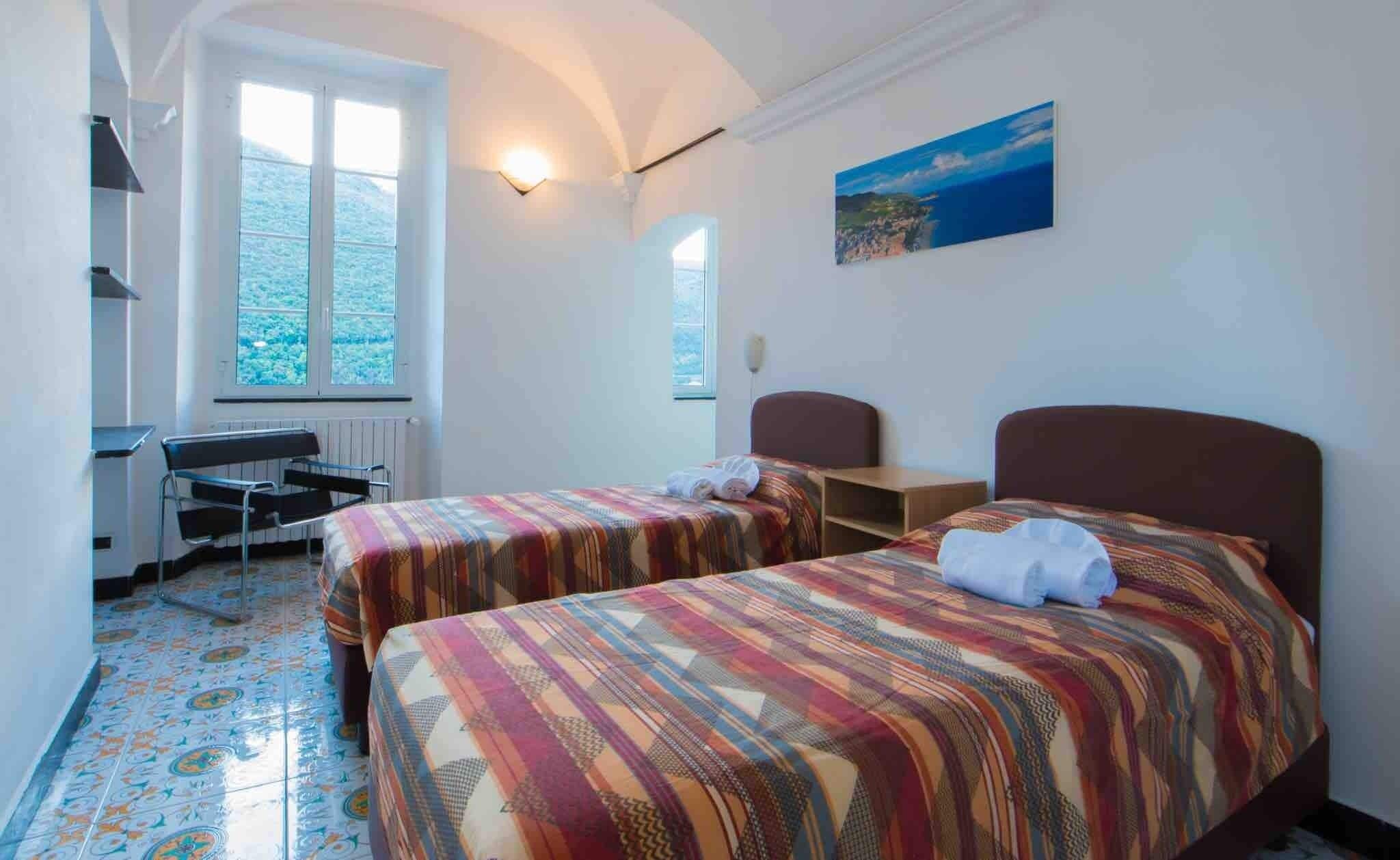 A room with two beds, a desk, and a chair. There are windows with a view of the sea.