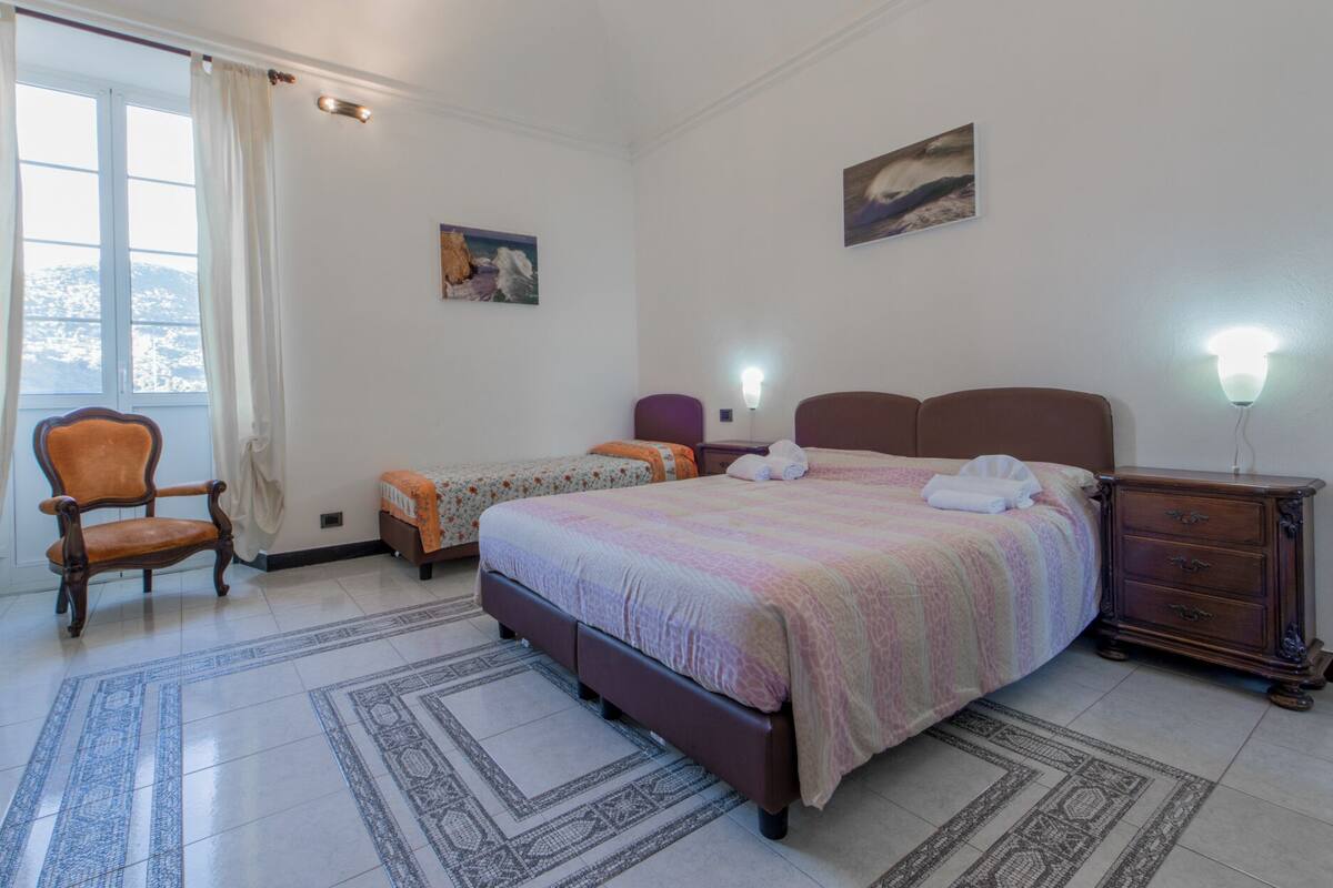 Ein Hotelzimmer mit einem großen Bett, zwei kleineren Betten, einem Sessel, einer Kommode und einem Fenster mit Vorhängen.