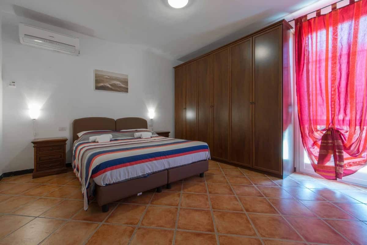 Ein Schlafzimmer mit Bett, Kleiderschrank, Nachttisch, Wand-Klimaanlage und Fenster mit roten Vorhängen.