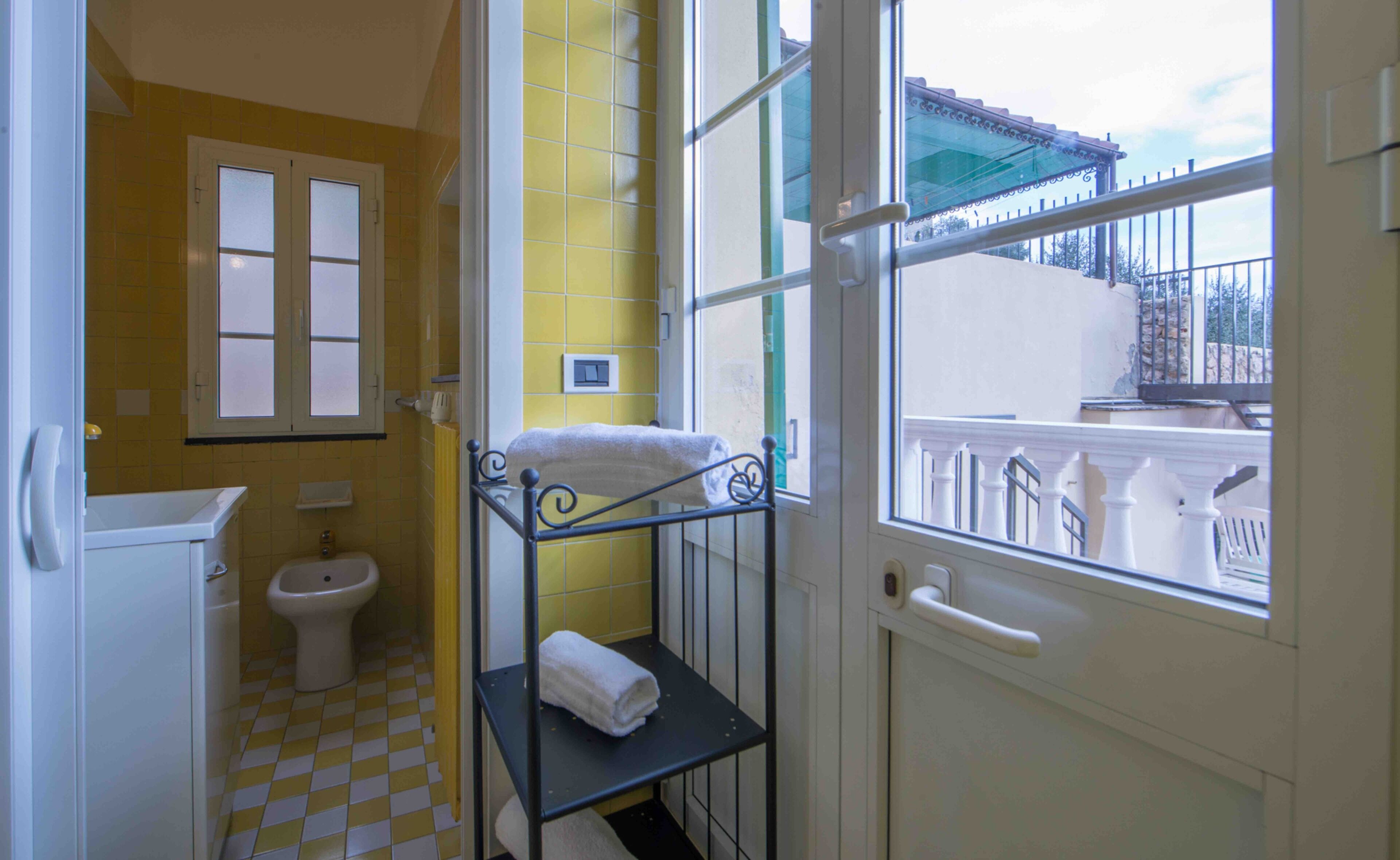 Une salle de bain avec des carreaux jaunes, des toilettes blanches, un lavabo et une fenêtre donnant sur un balcon.