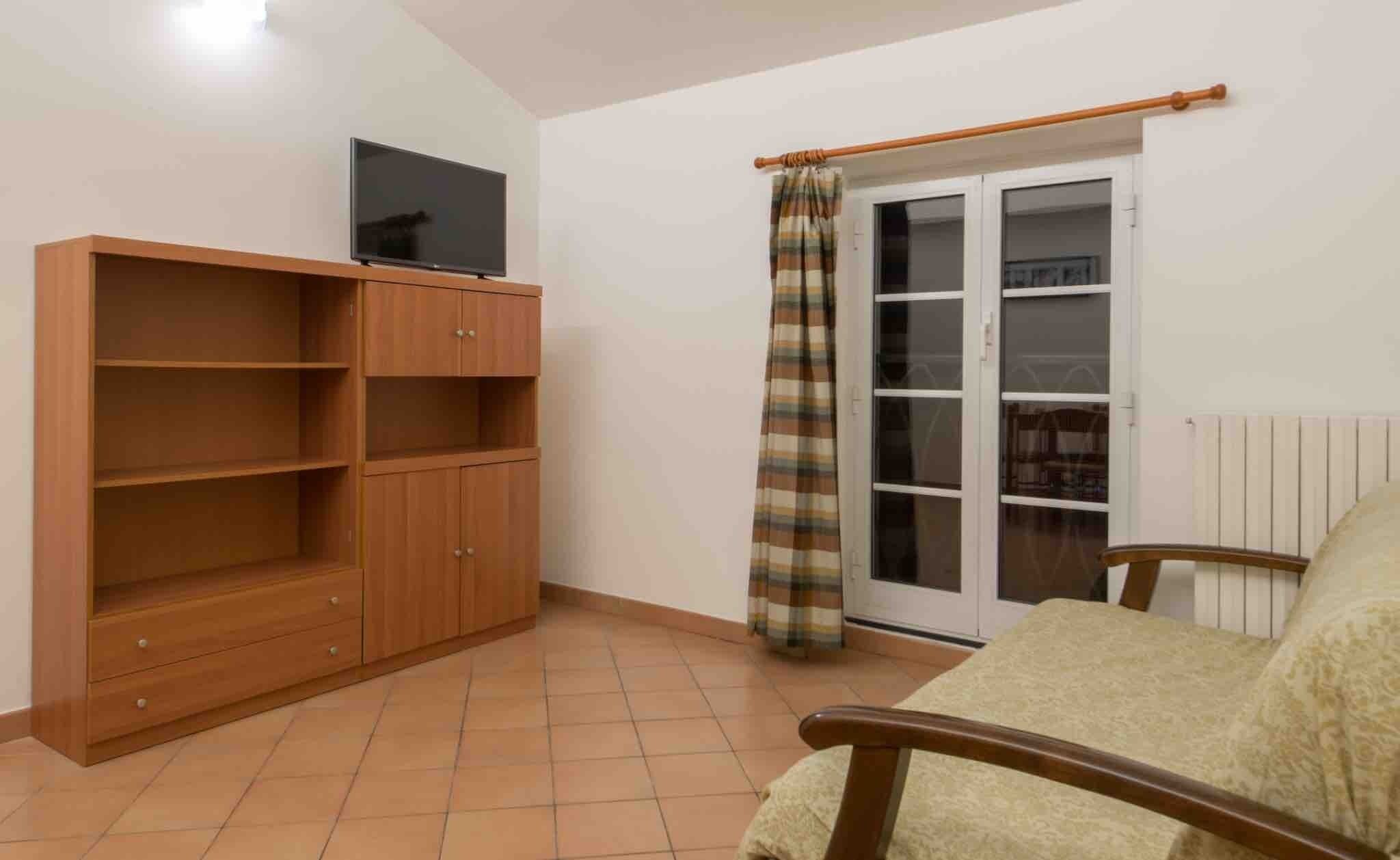 A room with a wooden cabinet, a television on top, a plaid curtain, a sliding glass door, and a beige armchair.