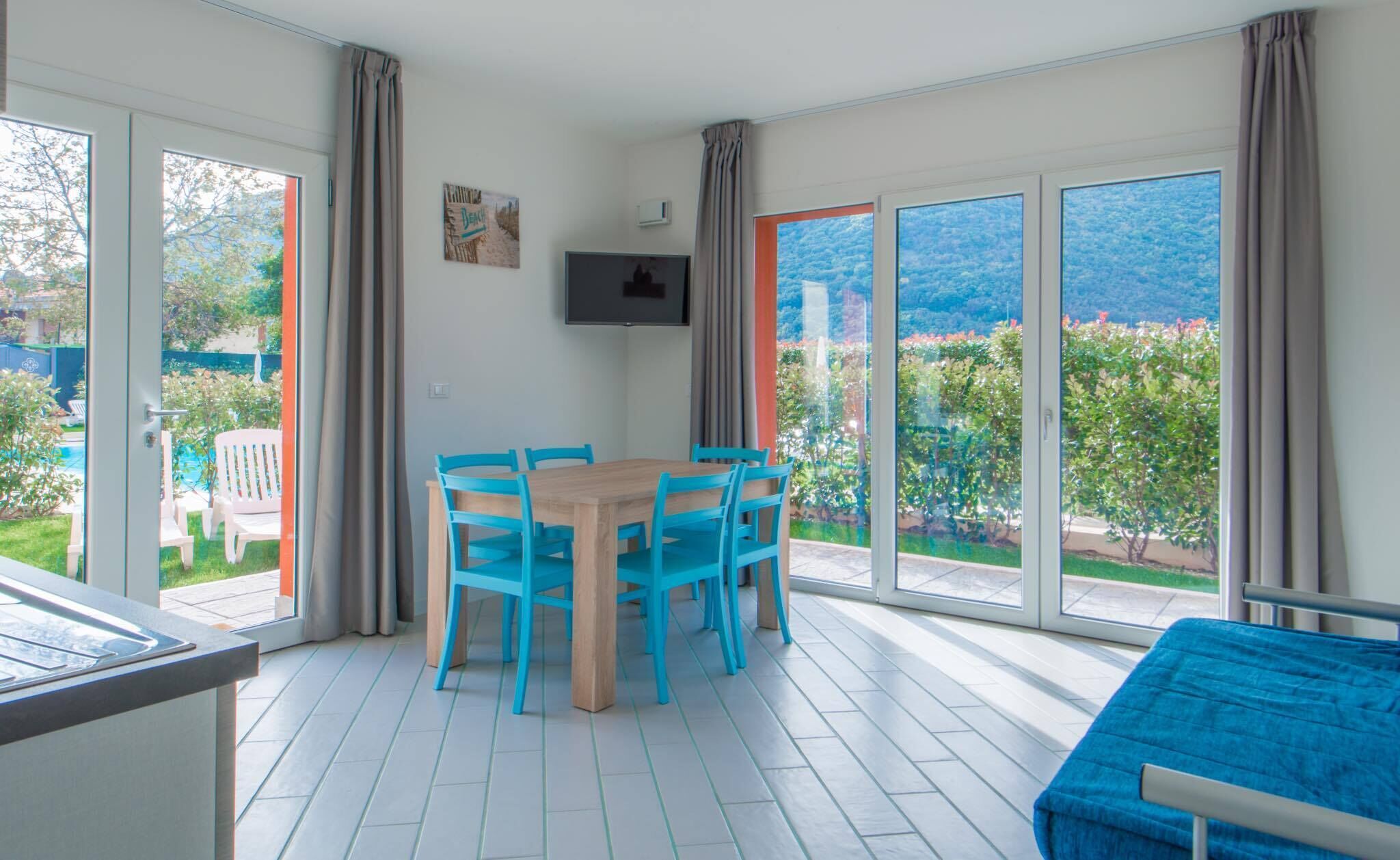 Ein modernes Zimmer mit Esstisch und Stühlen, einem an der Wand montierten Fernseher und großen Schiebeglastüren, die auf einen Balkon mit Blick auf Grünflächen und Berge führen.