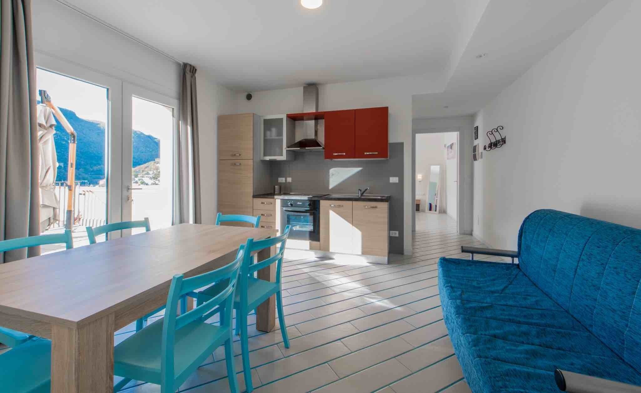 Una cucina moderna con mobili rossi, un'area pranzo con tavolo in legno e sedie color turchese, e un divano blu.