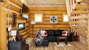 A log cabin interior with a black leather sofa, wooden walls, a fireplace, and a wooden staircase. - Custom Log Home directly along the Greenbrier River sits the River Ridge Retreat (Marlinton)