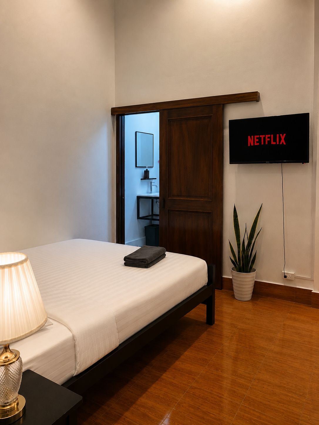 Ein Hotelzimmer mit einem Bett, einer Nachttischlampe, einem an der Wand montierten Fernseher und einer Zimmerpflanze.