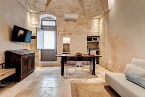 A stone-walled room with a wooden table, clear chairs, a sofa, a TV, and a kitchen area with a microwave.