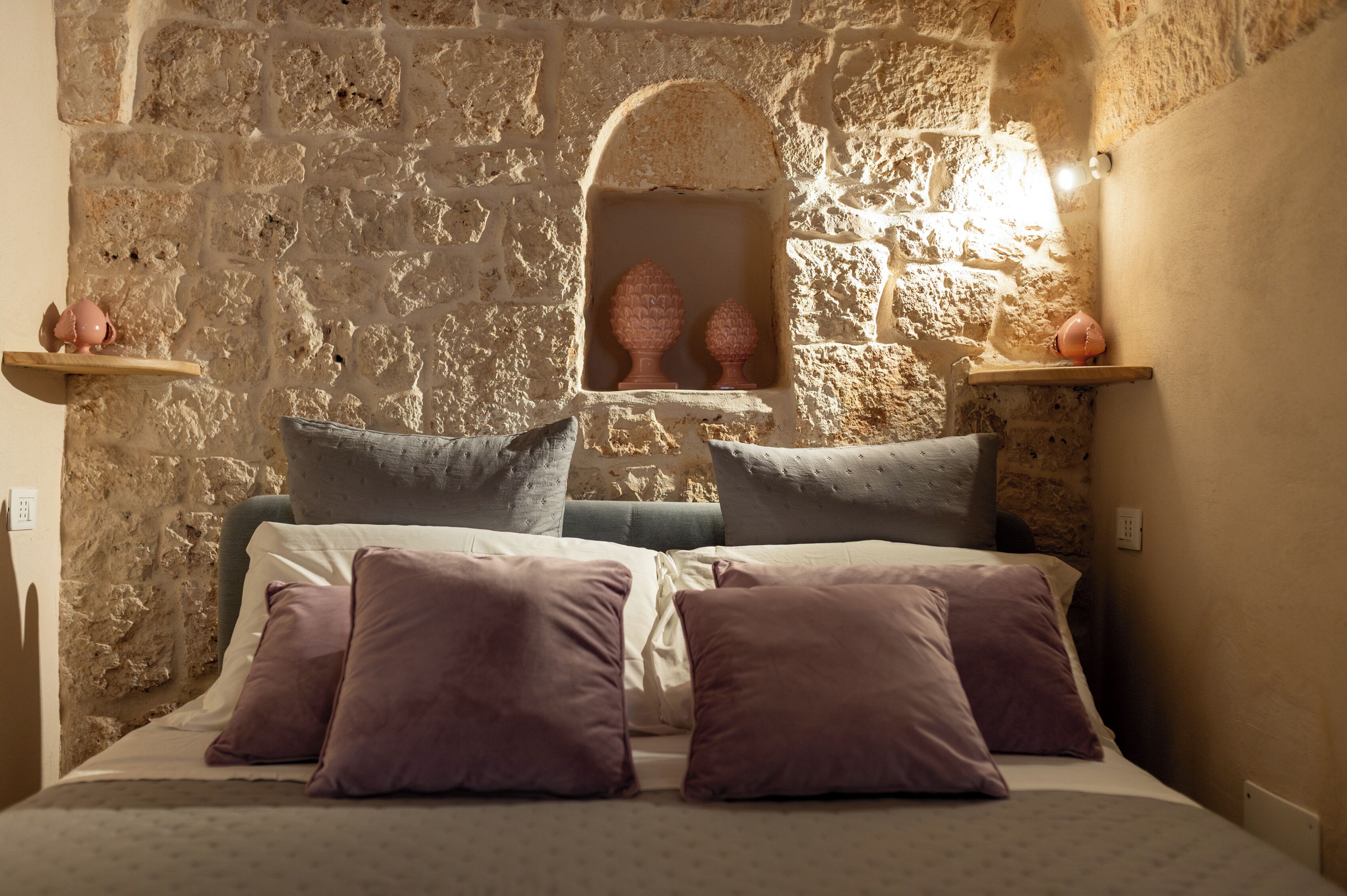 Ein Schlafzimmer mit Steinwänden, einem Bett, Kissen und einem kleinen Regal mit Dekoration.
