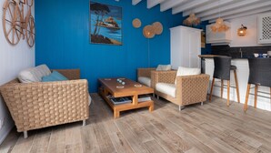 A room with wicker furniture, a wooden coffee table, a painting, and a kitchen area with bar stools.