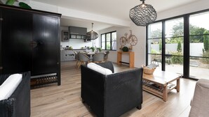 Ein modernes Wohnzimmer mit Rattanmöbeln, einem Holztisch und Blick auf die Küche und den Garten durch große Fenster.