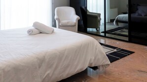 Ein modernes Hotelzimmer mit einem großen Bett, einem weißen Sessel, einem kleinen Tisch und einem großen Spiegel.