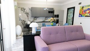 Ein modernes Wohnzimmer mit einem violetten Sofa, einem Essbereich mit Tisch und Stühlen, einer Küche mit Marmorarbeitsplatten und einem farbenfrohen, abstrakten Gemälde an der Wand.