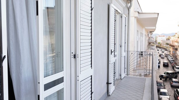 Ein schmaler Balkon mit Fliesenboden, weißen Fensterläden und einer Reihe geparkter Autos auf der rechten Seite.