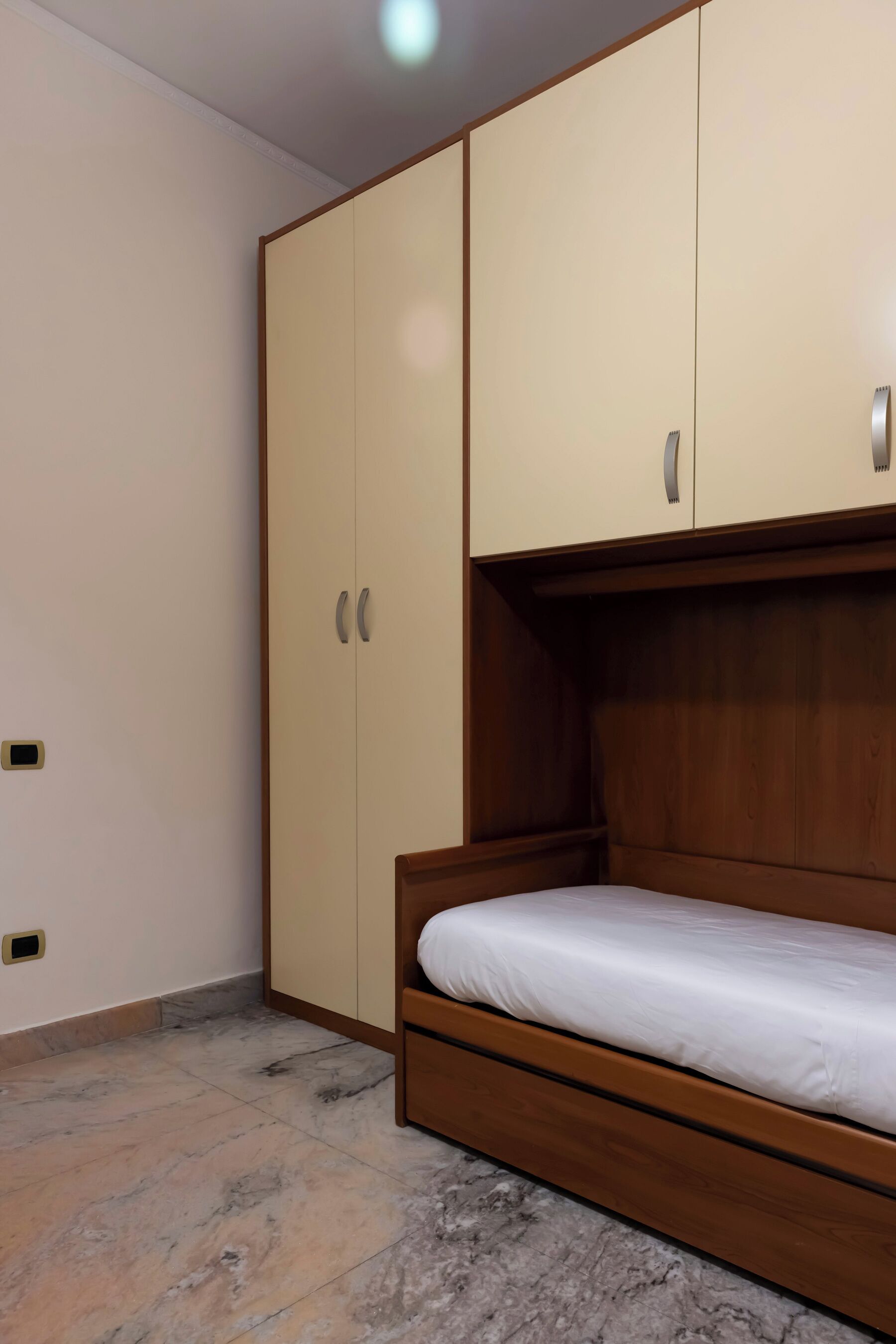 A bedroom with a wooden bed, a beige wardrobe, and a marble-tiled floor.