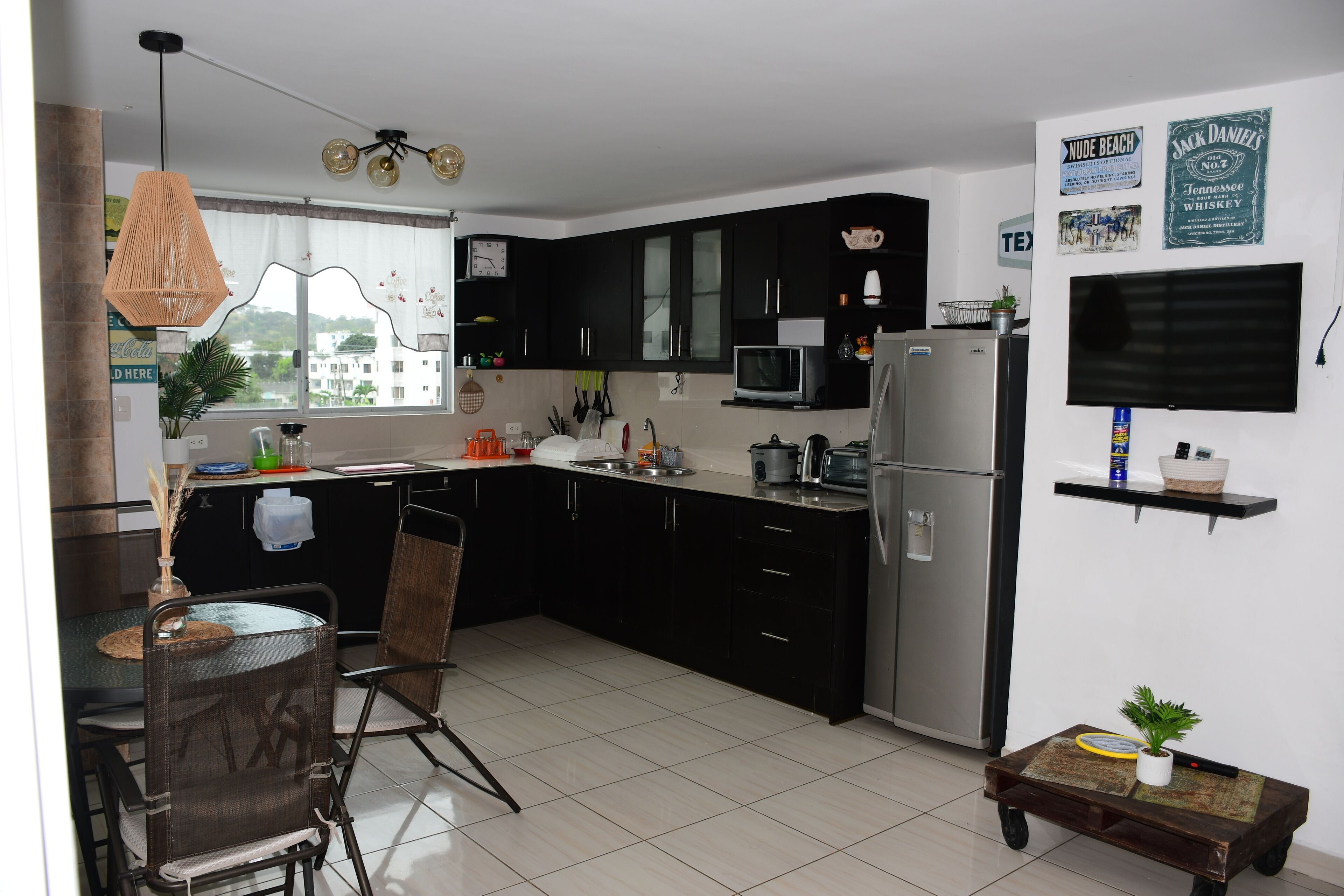 Una cucina moderna con mobili neri, un frigorifero in acciaio inossidabile, un forno a microonde e un televisore montato a parete. C'è un'area pranzo con tavolo e sedie, e un piccolo tavolino con una pianta.