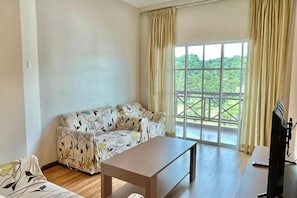 A living room with a sofa, a coffee table, and a TV. There is a large window with curtains. - PLAY at Iris Resort Tanah Rata (Cameron Highlands)