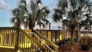 Eine Holztreppe führt zu einer Terrasse, im Hintergrund sind Palmen zu sehen.