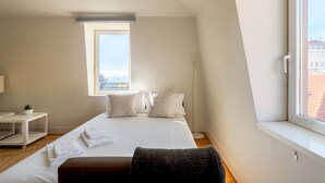 A bedroom with a large bed, a bedside table, a small plant, and a view of buildings through a window.