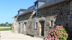 A stone building with multiple windows and a door, a gravel driveway, and a bush of pink flowers. - Gîte ti glas - Les Gîtes de la Cour Chevalier (La Fresnais)