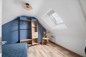 Ein Schlafzimmer mit schrägem Dach, einem Oberlicht, einem Kleiderschrank, einem Sessel und einem Bett mit gemusterten Betttextilien.