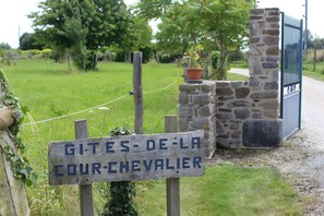 Paredão rústico de pedra com uma placa de madeira indicando 'GITES-DE-LA COUR-CHEVALIER' em frente a um portão de metal.