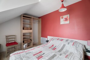 Ein Schlafzimmer mit schrägem Dach, einem Holzschrank, einem Bett mit gemustertem Überwurf, einer roten Wand und einer Pendelleuchte.
