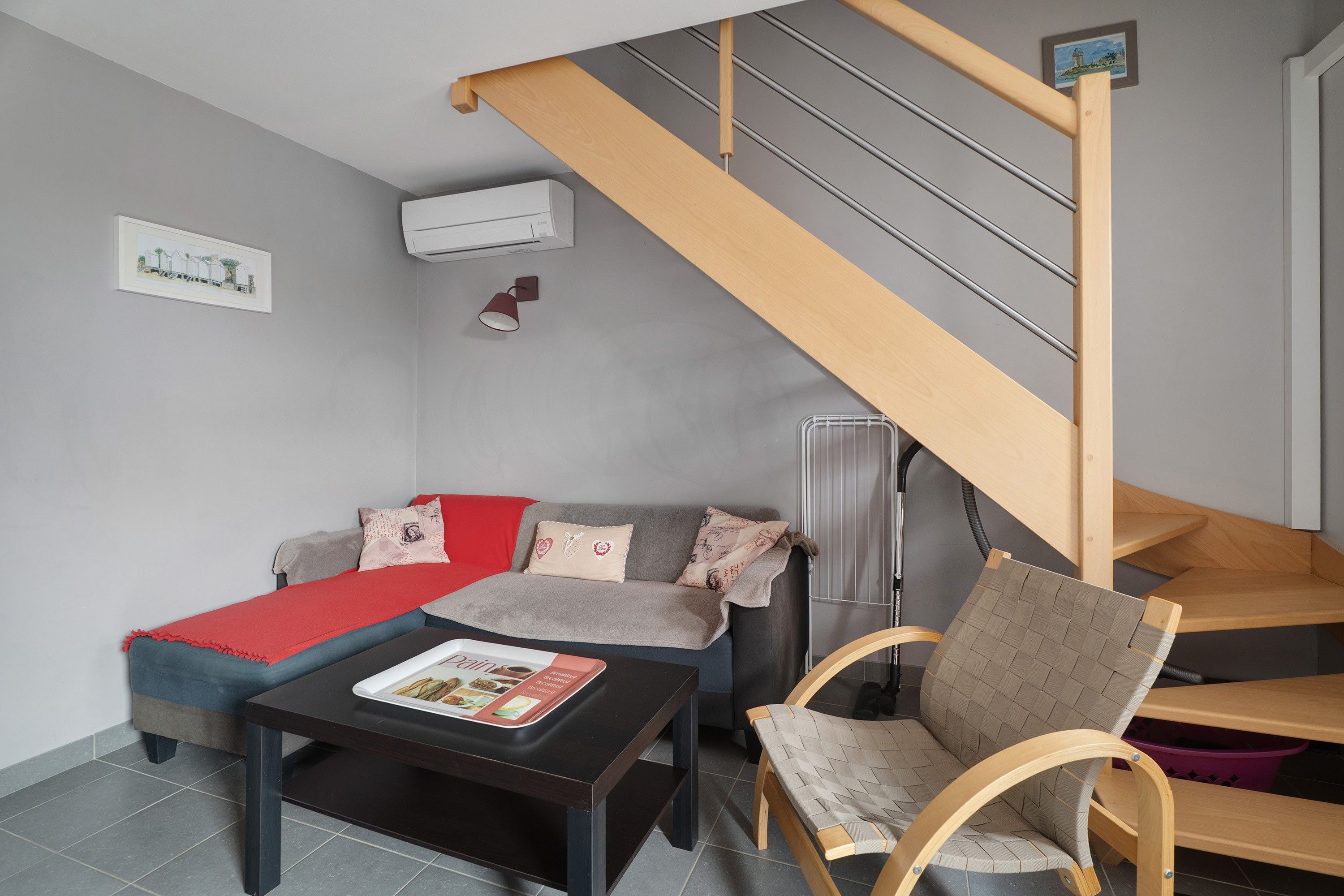 A modern living room with a sofa, a coffee table, and a wooden chair. A staircase with a wooden railing leads to an upper level.