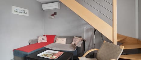 Una sala moderna con un sofá, una mesita de centro y una silla de madera. Una escalera con barandilla de madera conduce a un nivel superior.
