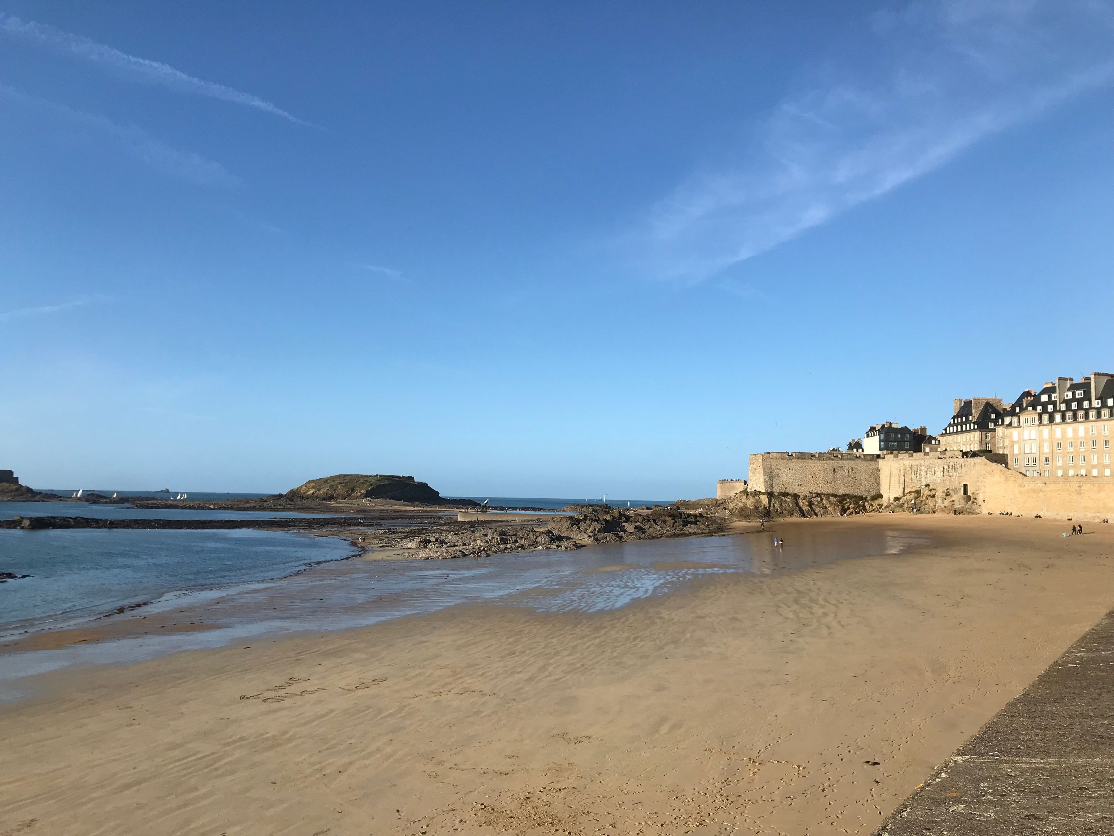 Una playa costera con un edificio histórico de piedra, costa rocosa y cielo azul despejado.