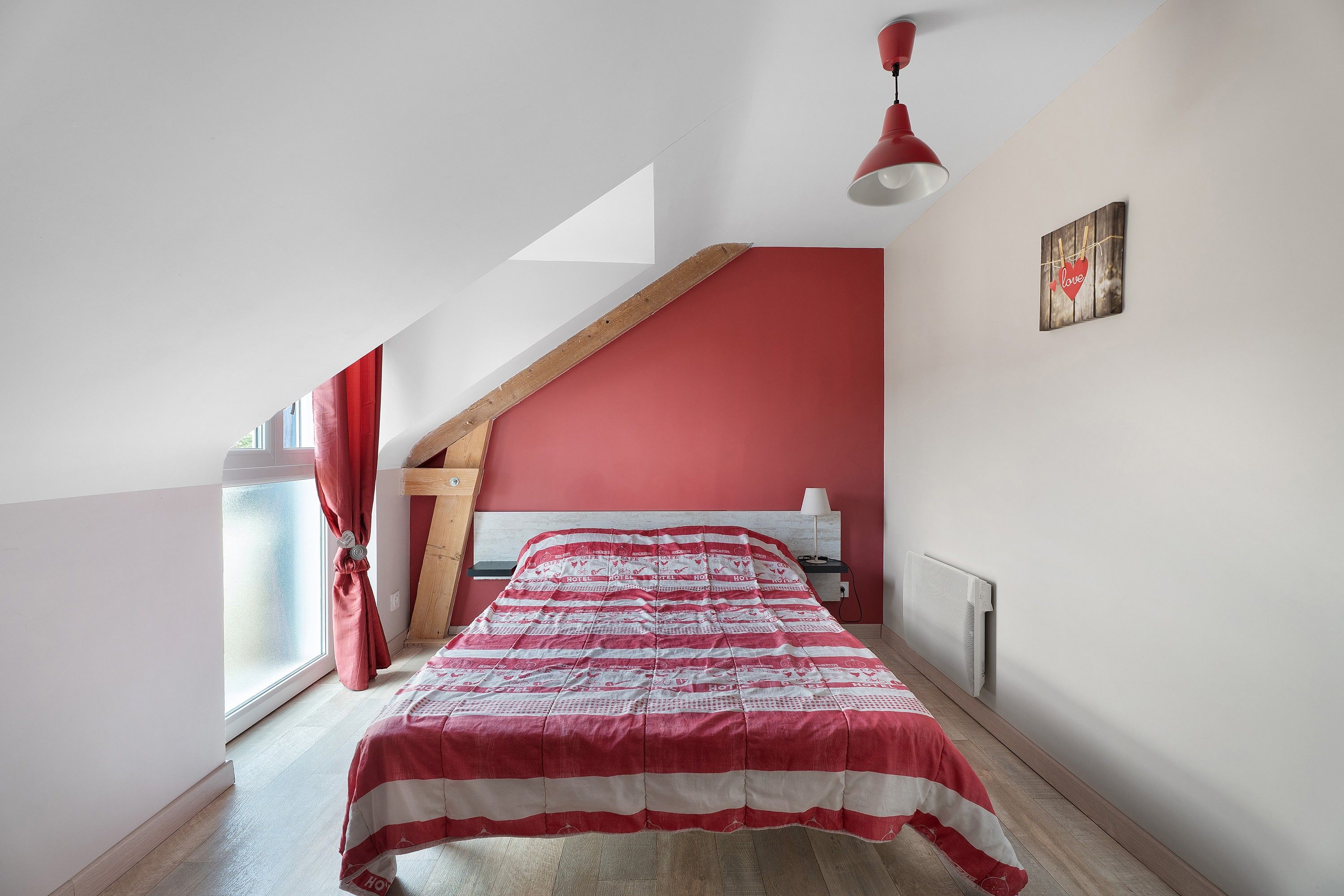 A bedroom with a sloped ceiling, a bed with a striped bedspread, a red wall, a window with red curtains, and a wall-mounted lamp.
