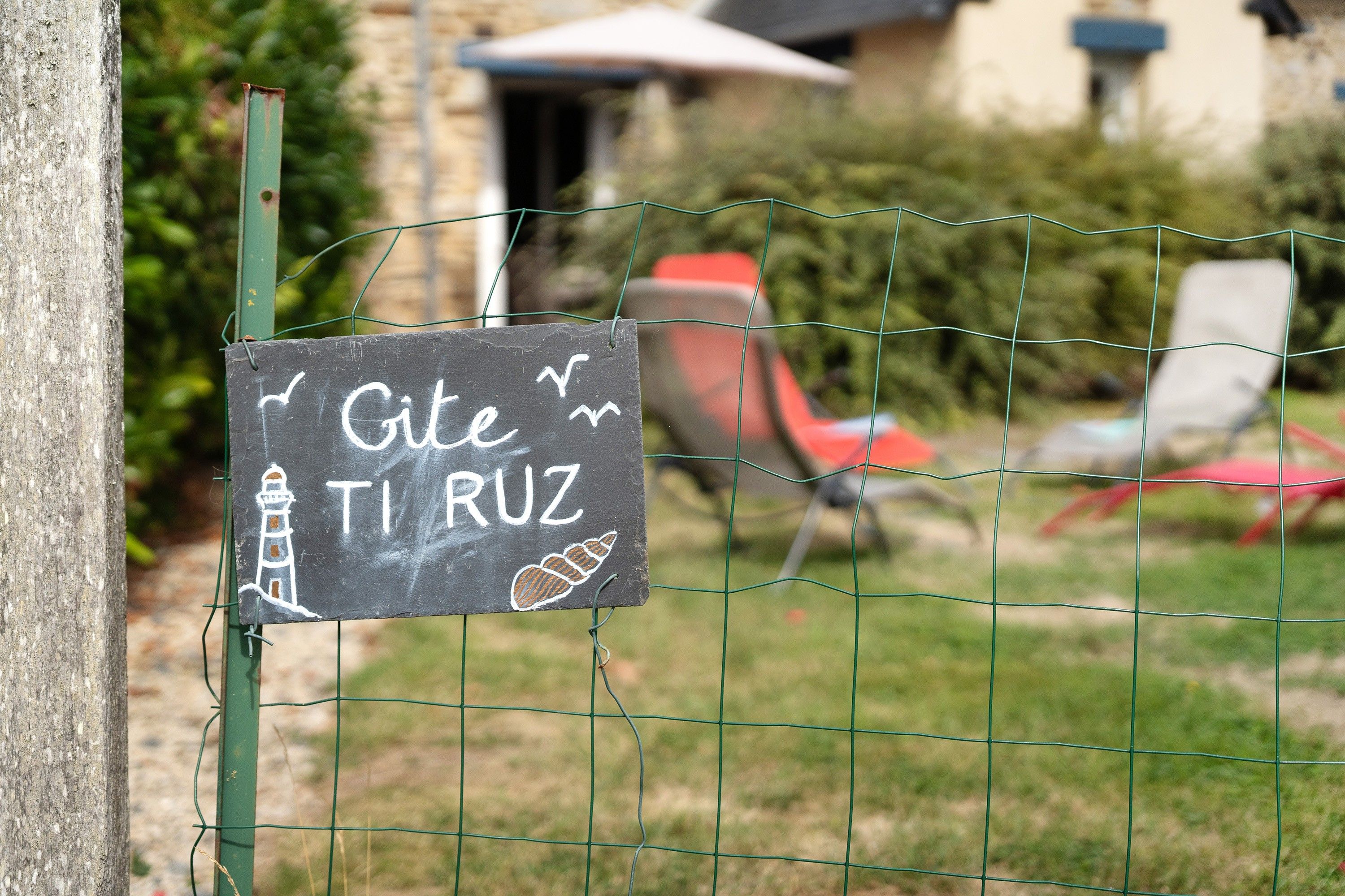 Ein Schild mit dem Schriftzug 'Gite TI RUZ' und einer Zeichnung eines Leuchtturms, an einem grünen Metallpfosten befestigt.