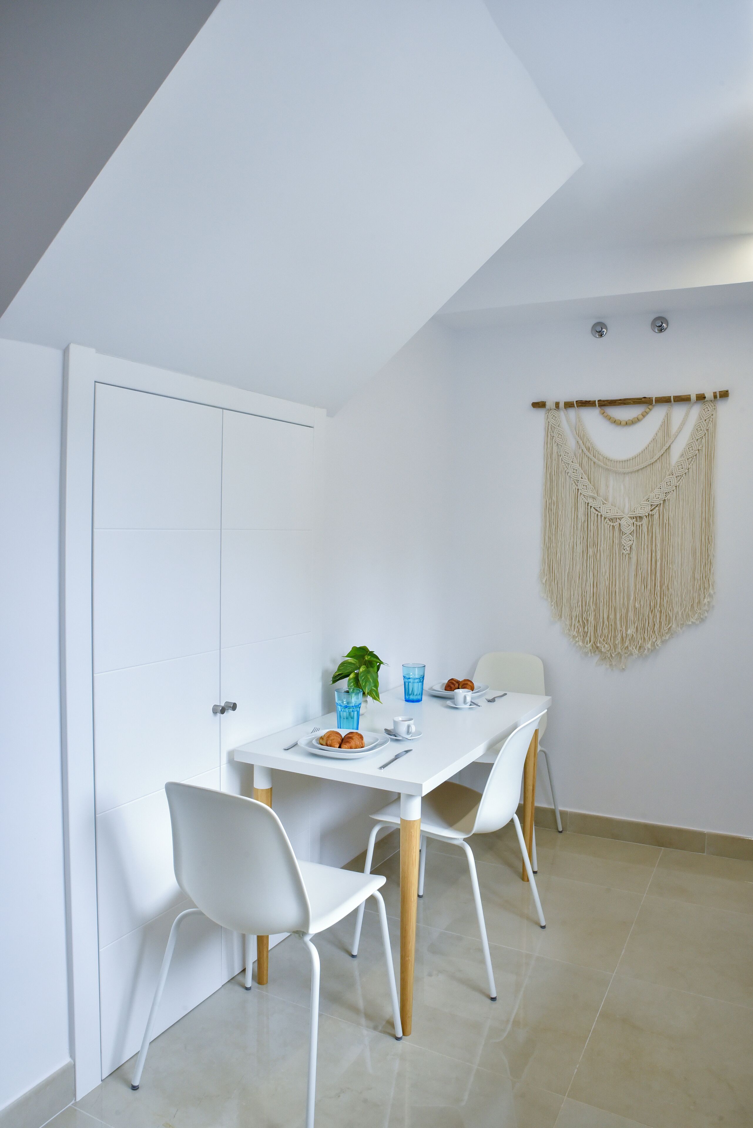 A modern dining area with a white table, four white chairs, a macrame wall hanging, and a door to the left.