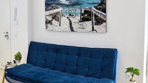 A modern living room with a blue sofa, a glass coffee table, and a wall-mounted painting of a coastal scene.