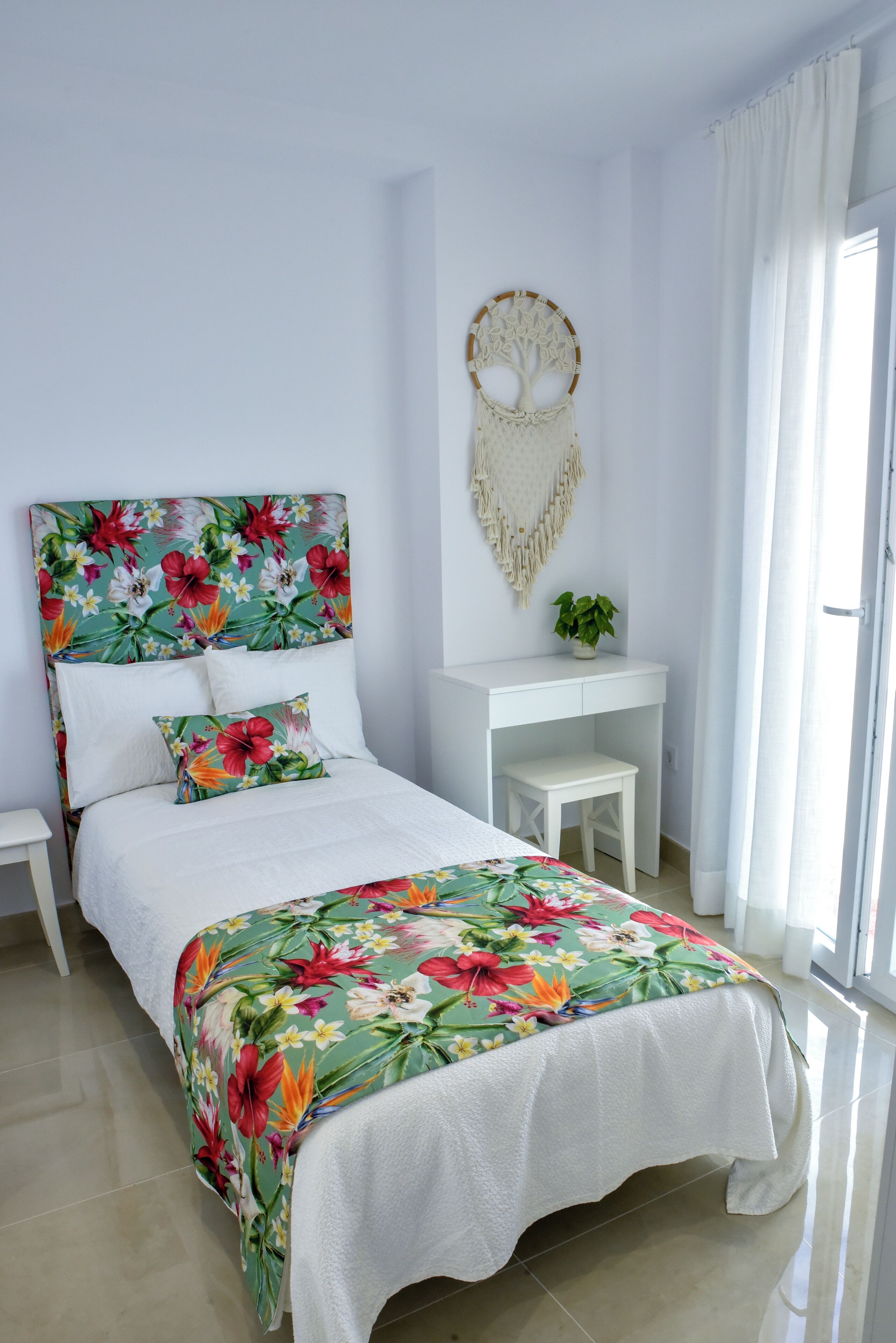 Un dormitorio con colcha estampada, cabecera blanca, una mesita de noche pequeña, escritorio blanco y un adorno en la pared.