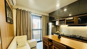 Eine moderne Küche mit Holztischplatte und eingebautem Herd. Ein weißes Sofa mit Kissen und ein kleiner Tisch mit Stühlen sind zu sehen. Ein Fenster mit Vorhängen ist erkennbar, und im Hintergrund steht ein Kühlschrank.