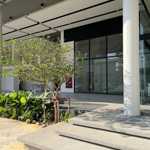 A modern building entrance with a glass facade, a small tree, and a concrete planter with various plants. - Hatyai Midtown Hotel (Hat Yai)
