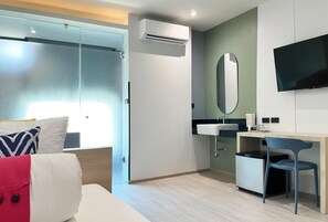 A hotel room with a bed, a desk, a chair, a mirror, a television, and an air conditioning unit.