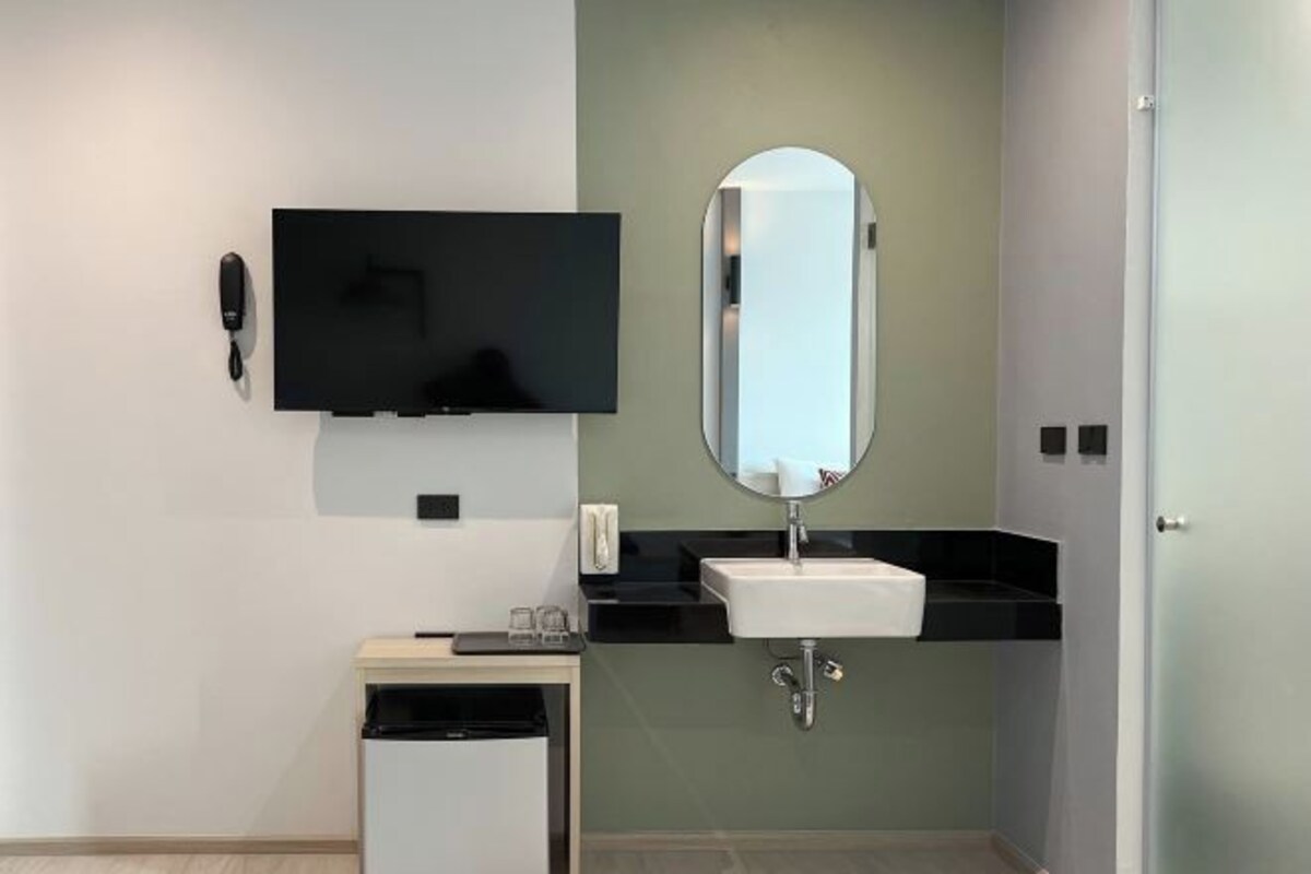 A modern bathroom with a flat-screen TV mounted on the wall, a sink with a mirror, and a small trash bin.