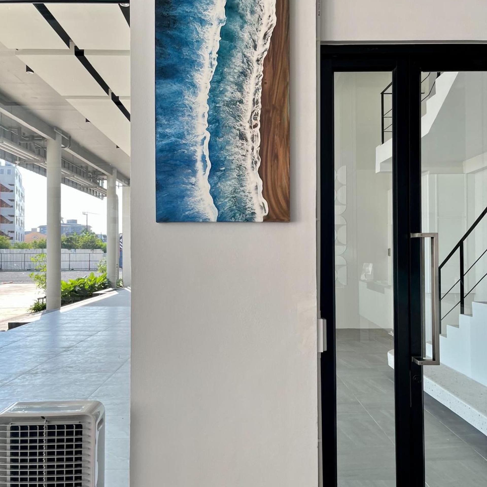 An interior corridor with a large painting of ocean waves on the wall, a modern staircase, and a glass door leading to another area.