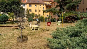 Un terrain de jeux avec des balançoires et un banc, entouré d’herbe sèche et d’arbres, avec des bâtiments en arrière-plan.