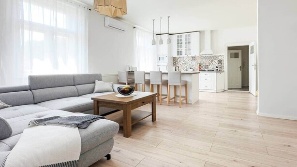 Un salon moderne avec un canapé gris, une table basse en bois et un coin cuisine doté de meubles blancs et d’un dosseret carrelé.