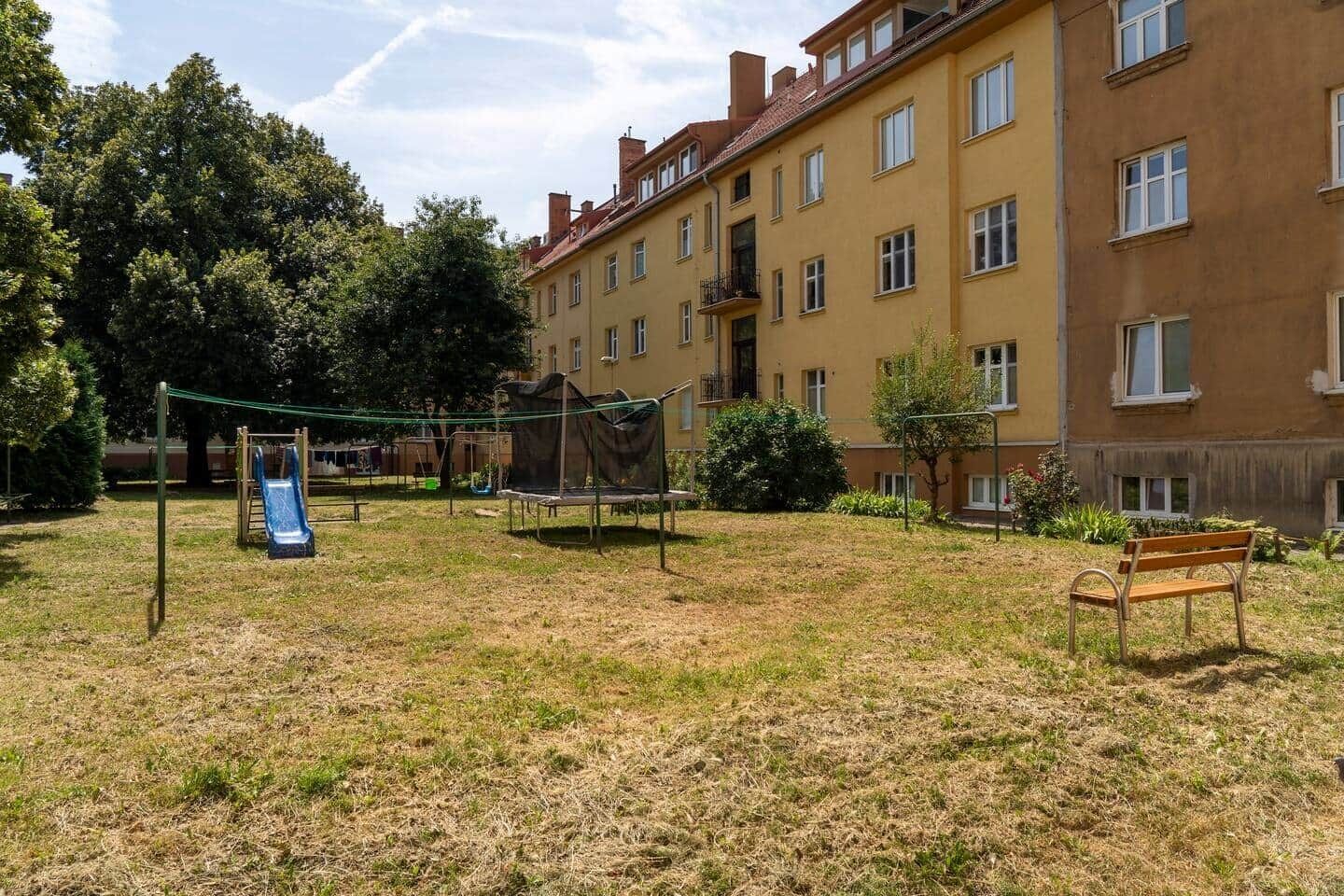 Un'area giochi con scivolo e trampolino, circondata da prati e alberi, con un edificio sullo sfondo.