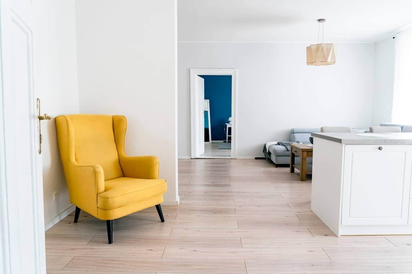 A modern living room with a yellow armchair, a wooden dining table, and a kitchen area with white cabinets.