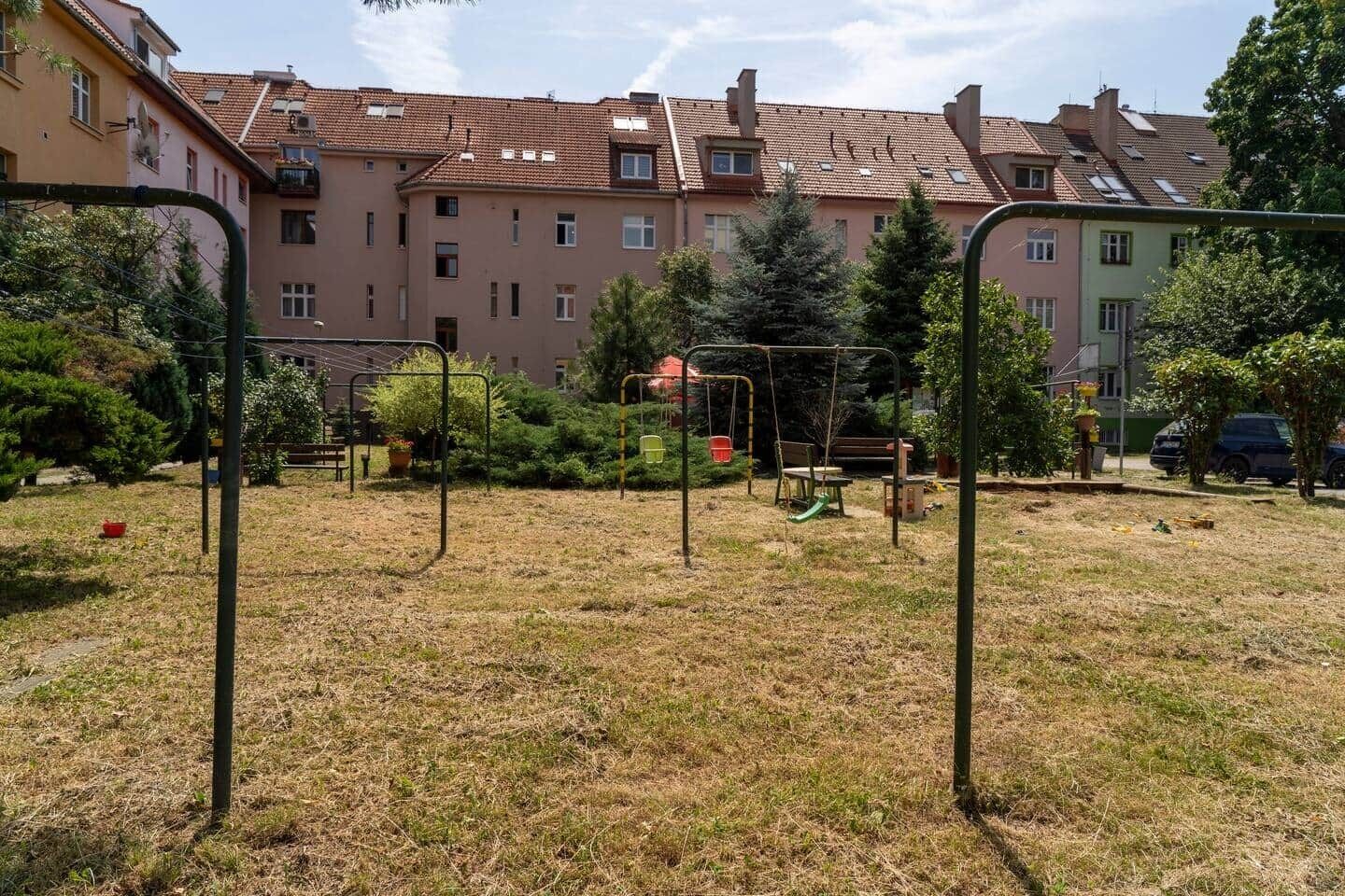 A playground with swings and a slide, surrounded by grass and trees, with apartment buildings in the background.