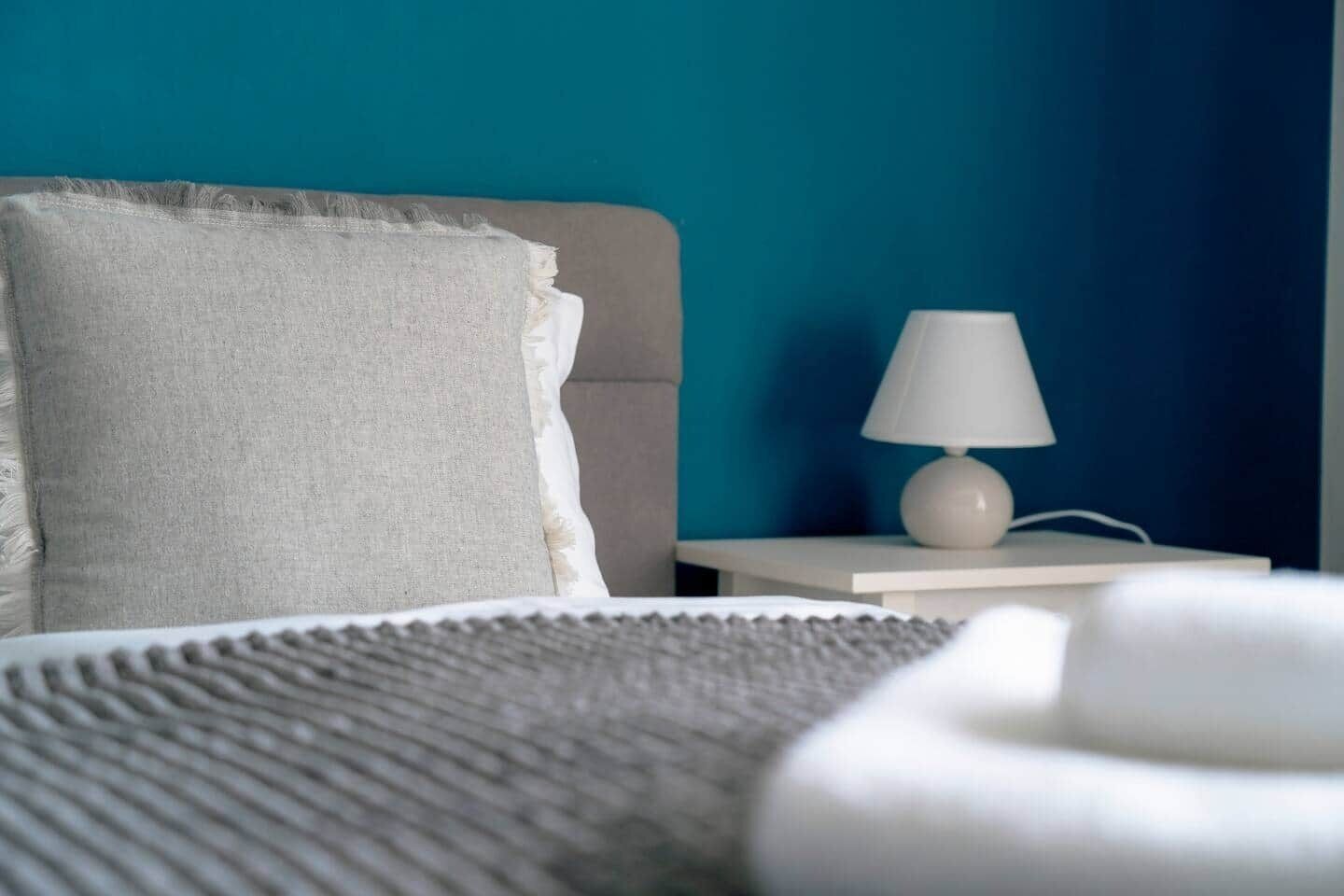 Un lit avec un oreiller blanc et une couverture grise, une table de chevet blanche avec une lampe, et un mur de couleur turquoise.