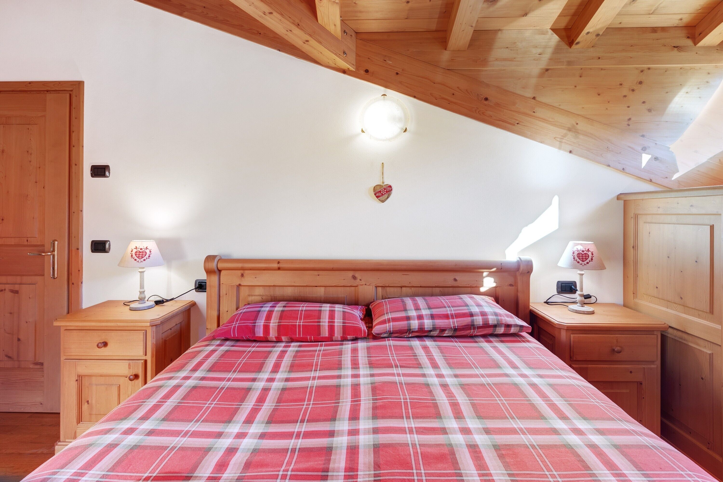 Una camera da letto con soffitto in legno, un letto con biancheria a quadretti, due comodini con lampade e un armadio in legno.