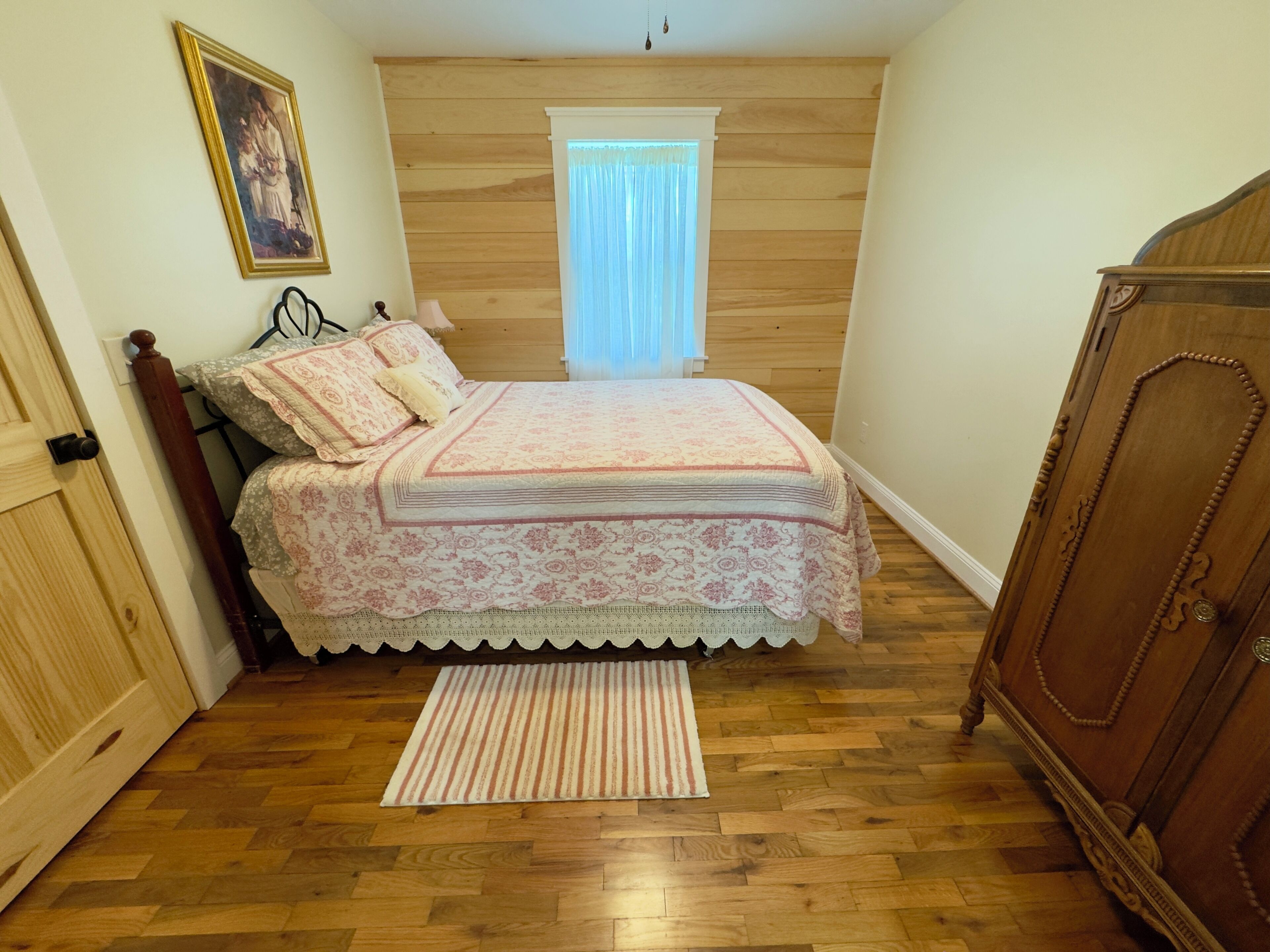A bedroom with a wooden floor, a bed with a patterned bedspread, a window with curtains, and a wooden wardrobe.