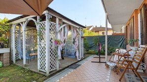 Un área de patio techada con una terraza de madera, una estructura de celosía blanca y mobiliario de exterior.