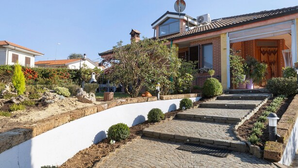 Propiedad residencial con camino de entrada pavimentado, antena parabólica, chimenea y jardín con diversas plantas y un pequeño árbol.