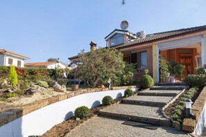 Propiedad residencial con camino de entrada pavimentado, antena parabólica, chimenea y jardín con diversas plantas y un pequeño árbol.
