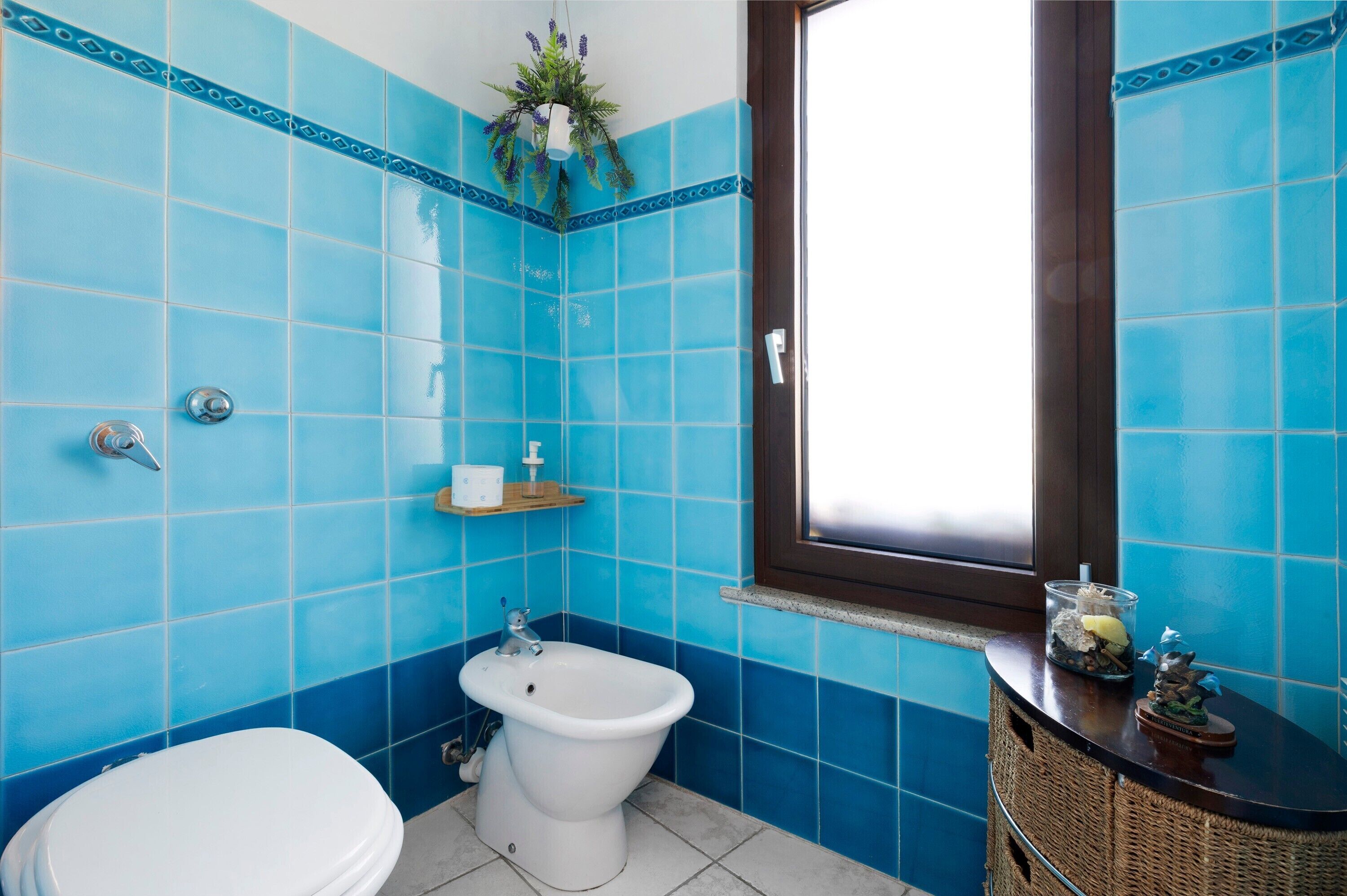A bathroom with blue tiles, a white toilet, a bidet, a small shelf with toiletries, and a window with a view.